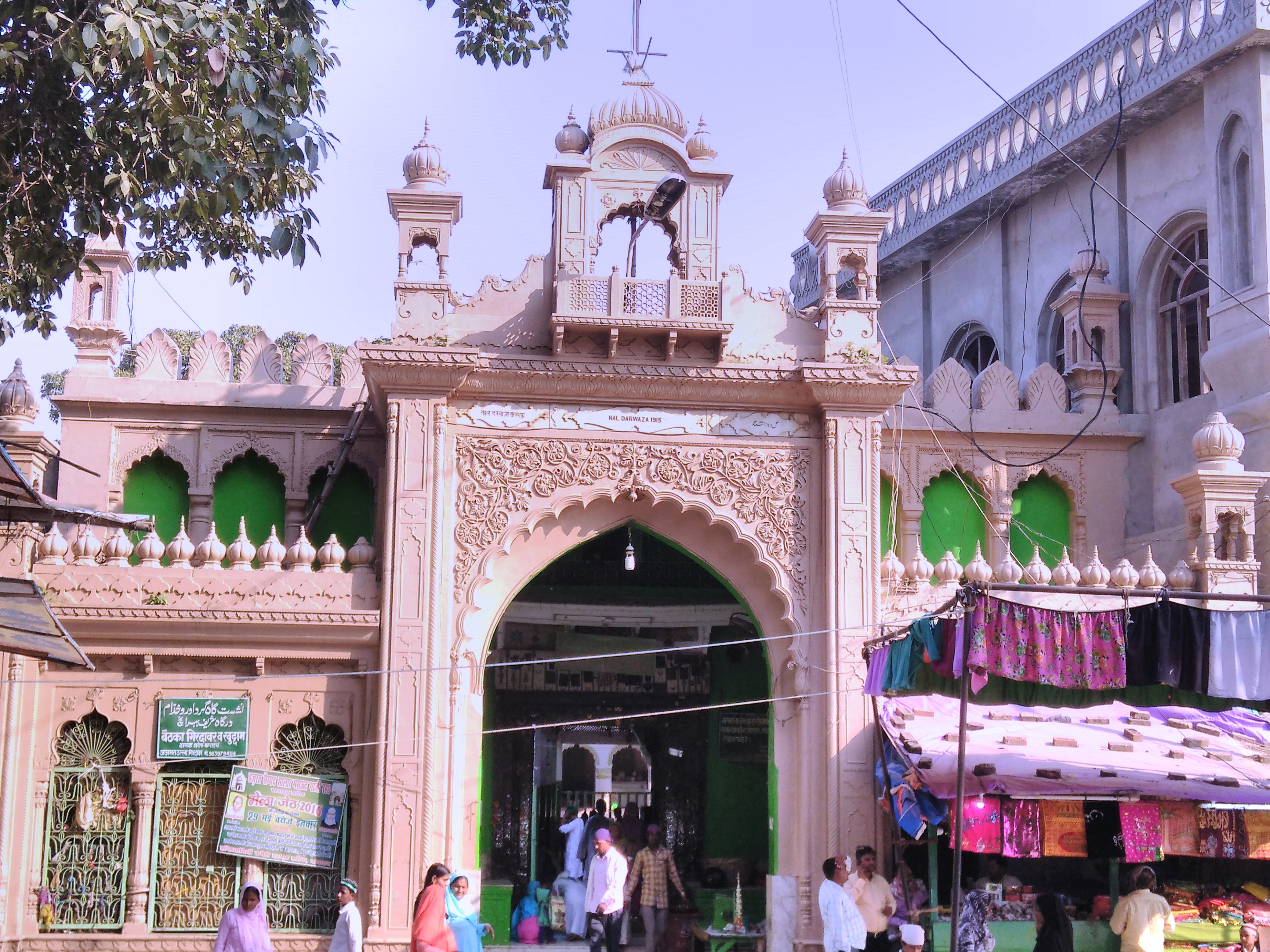 The large cream-colored gate of Salār Mas‘ūd’s shrine. People walk in, and there is a small shop in front